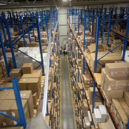 OSW Pro - Wysokie regały magazynowe wypełnione kartonami, oświetlone lampą. Widok z góry, z ręką w rękawicy trzymającą rurę. Perspektywa w głąb magazynu.