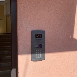 OSW Pro - Szary panel domofonu z klawiaturą i czytnikiem kart zbliżeniowych zamontowany na różowej ścianie budynku, obok ciemnych drzwi wejściowych.