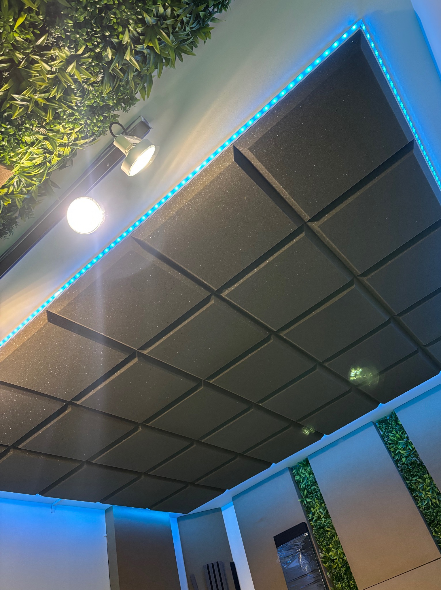 Sufit z paneli akustycznych w geometryczny wzór, podświetlony niebieską listwą LED, z elementami dekoracji roślinnej. Nowoczesny design wnętrza.