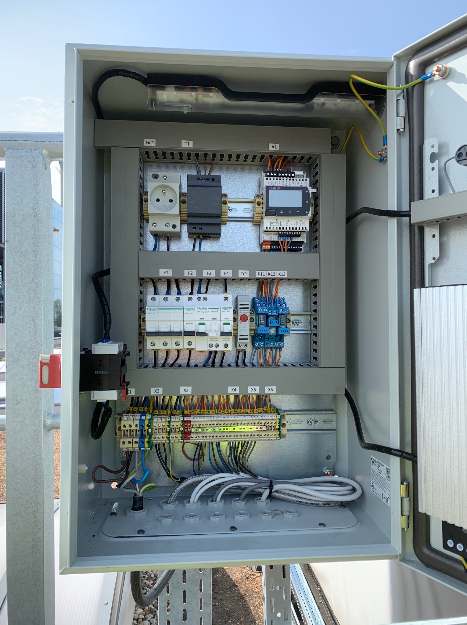 Otwarta, szara szafa sterownicza z widocznymi elementami elektrycznymi: gniazdo, bezpieczniki, przekaźniki, listwy zaciskowe i przewody. Widok z bliska na instalację.