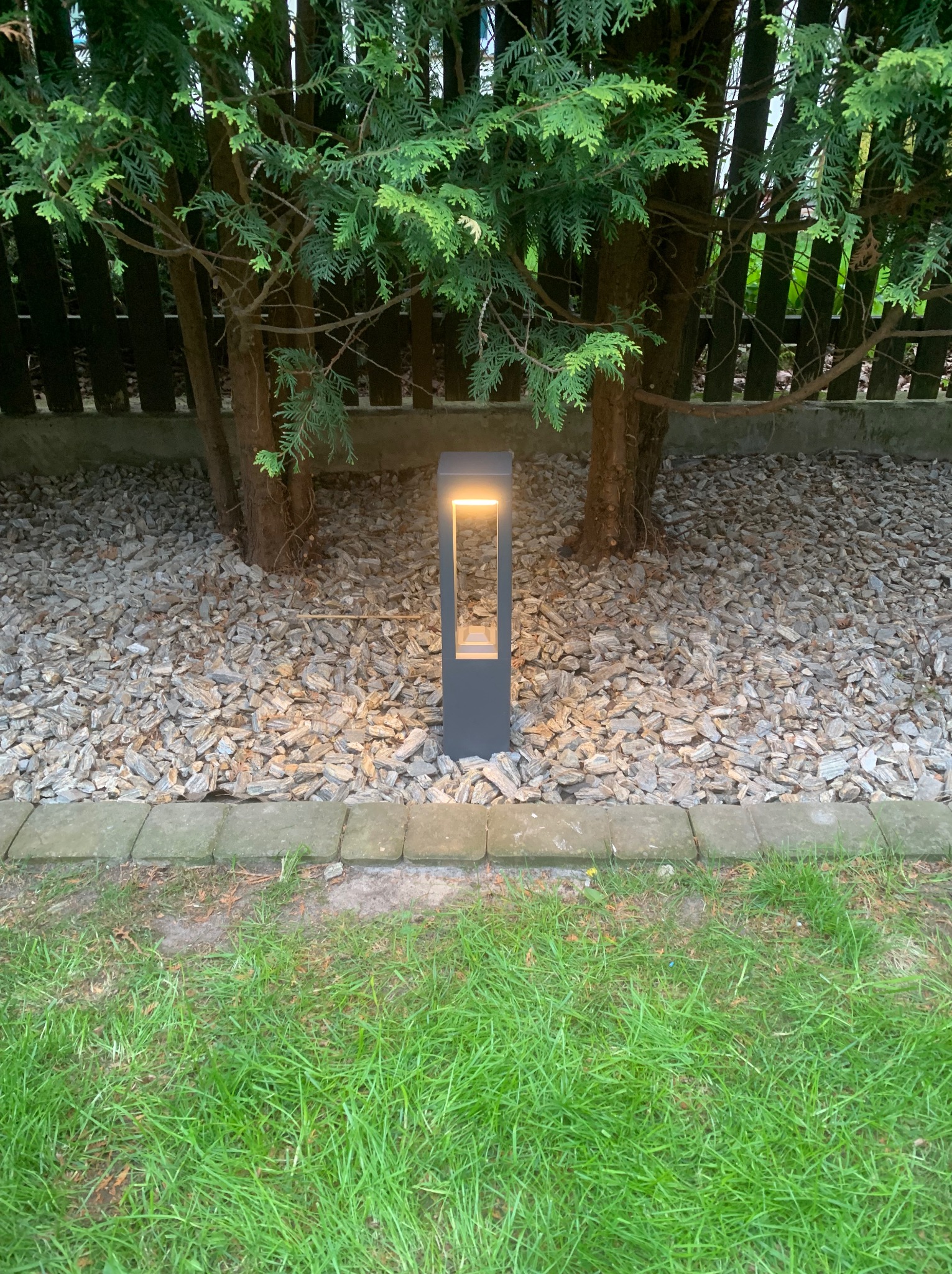 Nowoczesna lampa ogrodowa w kształcie prostopadłościanu, oświetlająca rabatę z korą drzewną i zielony trawnik. Oświetlenie zewnętrzne wkomponowane w krajobraz.