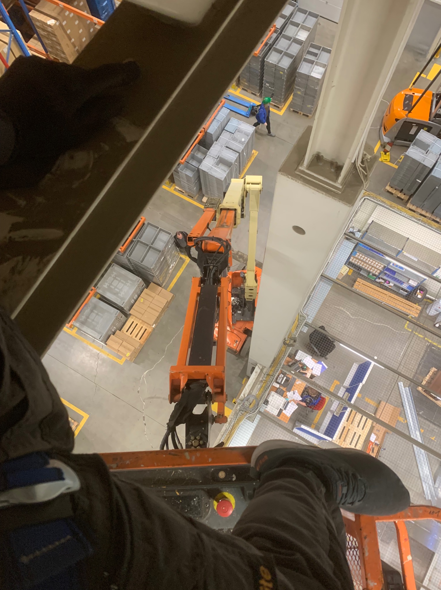 Widok z podnośnika koszowego na halę produkcyjną podczas prac na wysokościach. Widoczny pracownik w oddali i elementy konstrukcyjne hali.