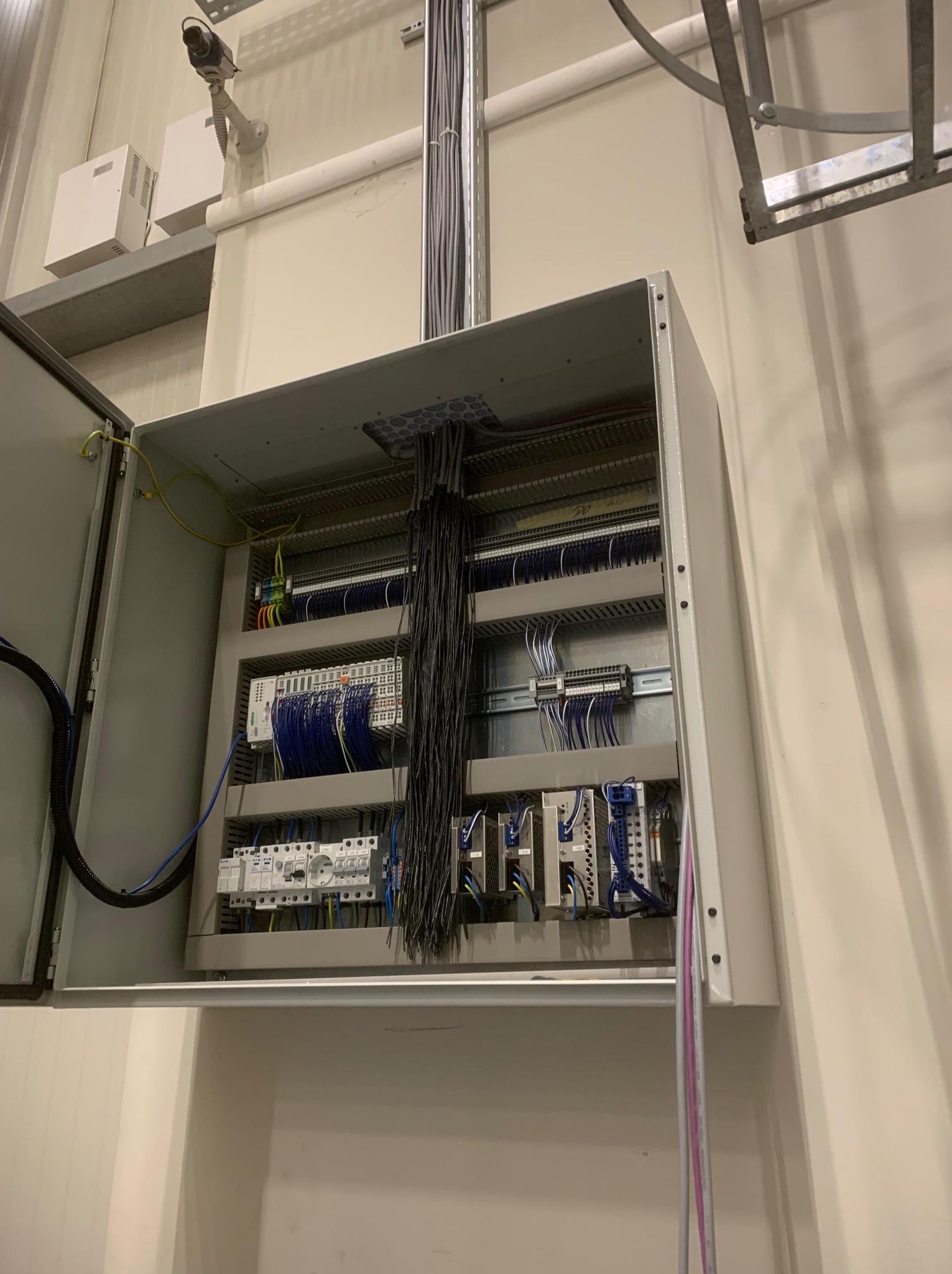 Otwarta szafa sterownicza z widocznymi elementami instalacji elektrycznej, przewodami i zabezpieczeniami. Widoczna kamera przemysłowa i kable w peszlu.