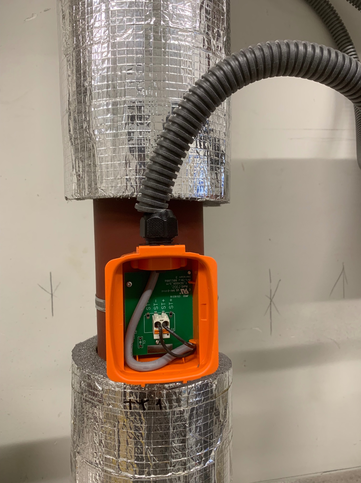 Pomarańczowa puszka instalacyjna z zieloną płytką drukowaną, podłączona do szarego przewodu karbowanego, zamontowana na rurze z izolacją termiczną w srebrnej folii.