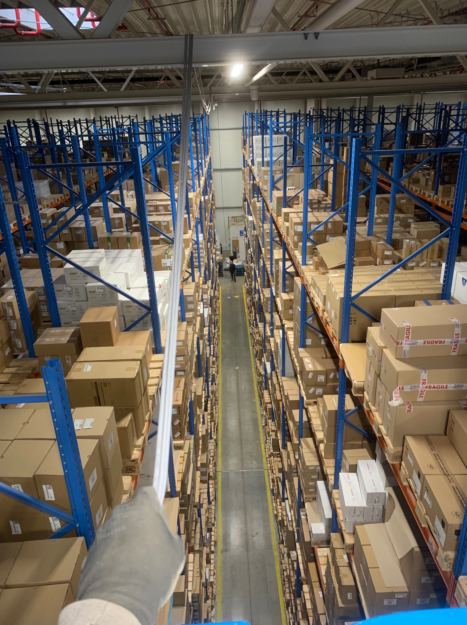 Wysokie regały magazynowe wypełnione kartonami, oświetlone lampą. Widok z góry, z ręką w rękawicy trzymającą rurę. Perspektywa w głąb magazynu.