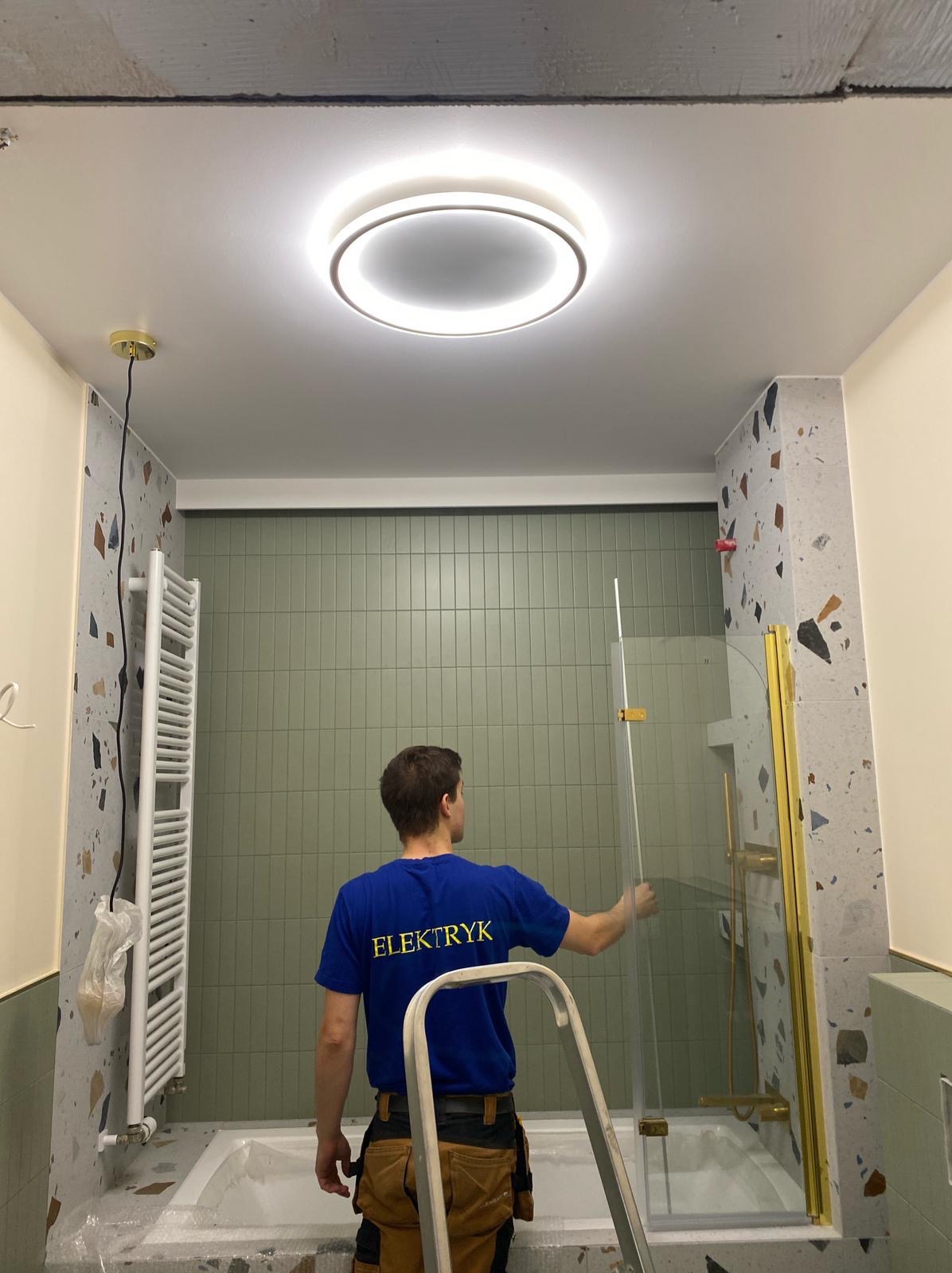 Elektryk montuje szklaną kabinę prysznicową w łazience z terazzo i zielonymi płytkami. Widoczne okrągłe oświetlenie sufitowe i drabina.