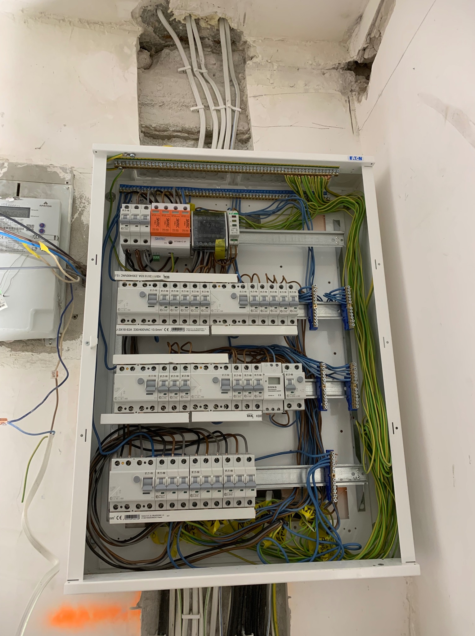 Rozdzielnica elektryczna w trakcie montażu w surowym wnętrzu, widoczne kable i zabezpieczenia. Częściowo wykończone ściany, instalacja w trakcie prac.