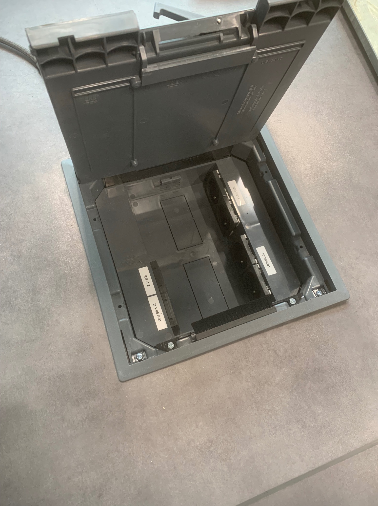 Otwarta, szara podłogowa puszka elektryczna z widocznymi gniazdami i oznaczeniami identyfikacyjnymi na etykietach, umieszczona na szarej podłodze.