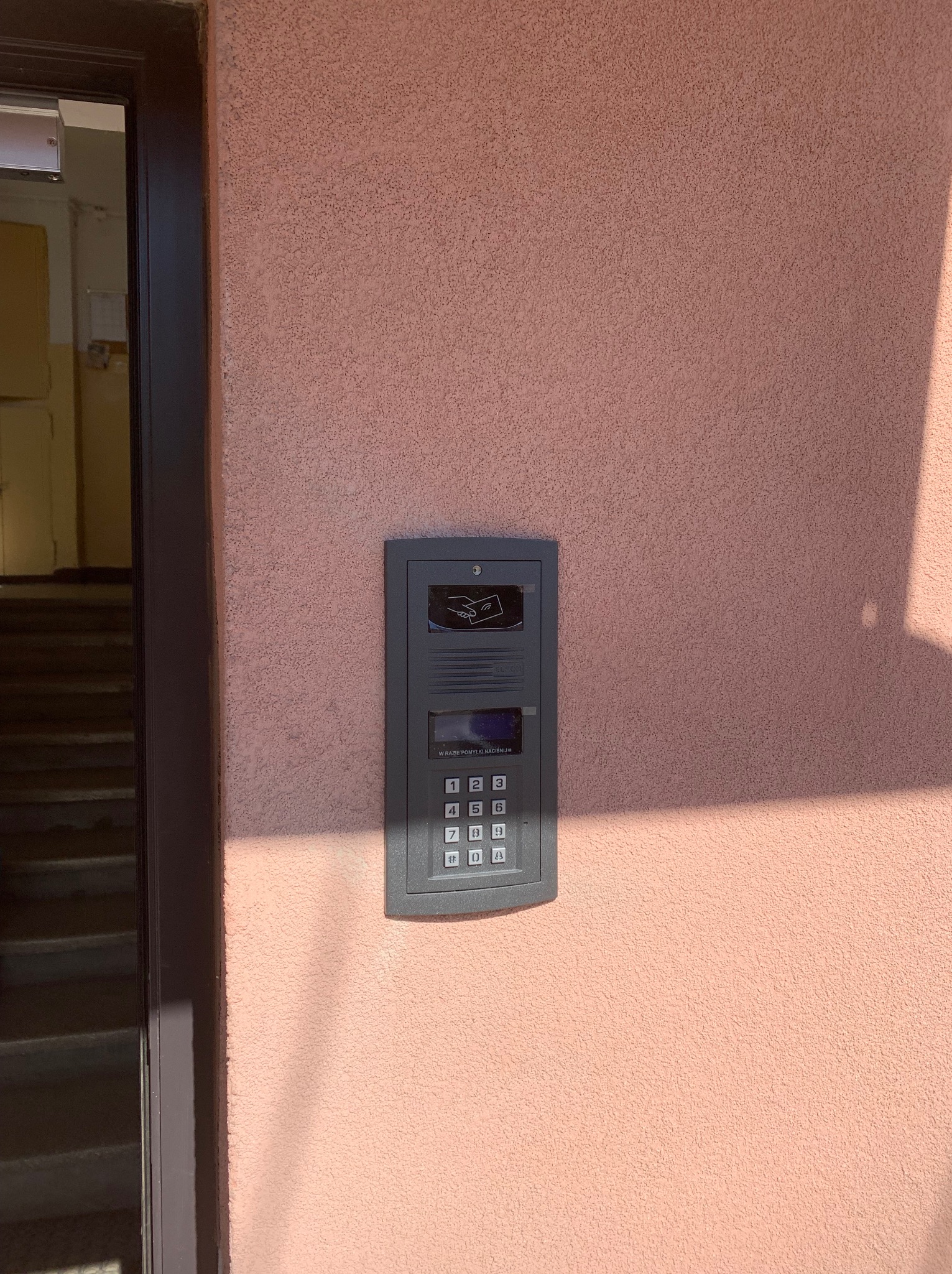 Szary panel domofonu z klawiaturą i czytnikiem kart zbliżeniowych zamontowany na różowej ścianie budynku, obok ciemnych drzwi wejściowych.