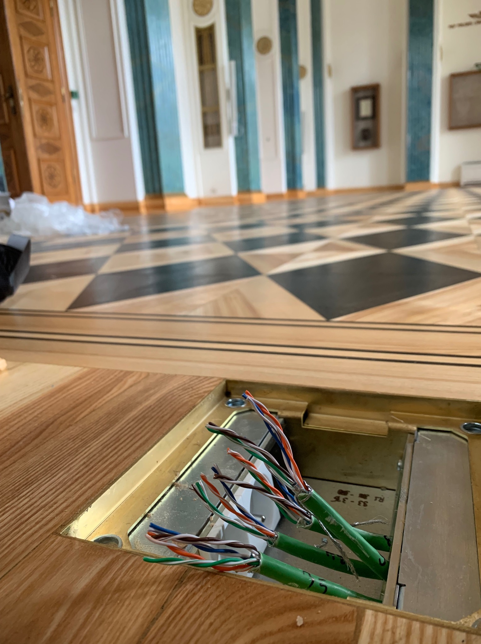 Podłączenie kabli w podłodze z parkietu w eleganckim wnętrzu. Widoczne kable telekomunikacyjne w metalowej puszce podłogowej. W tle wzorzysta podłoga i kolumny.