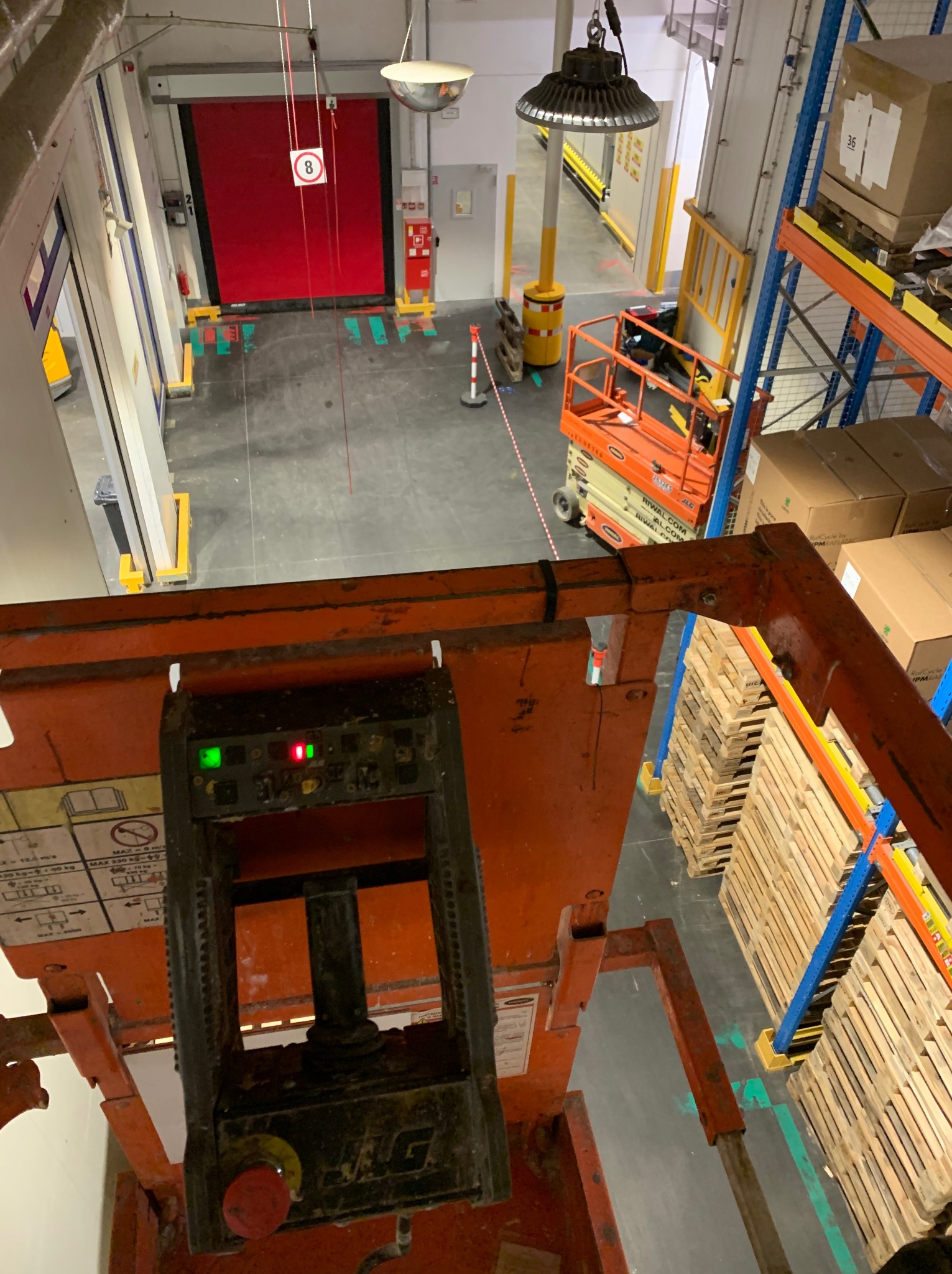 Widok z podnośnika koszowego na magazyn z regałami i bramą. W tle widoczne oświetlenie przemysłowe. Perspektywa z góry, z kabiny operatora.