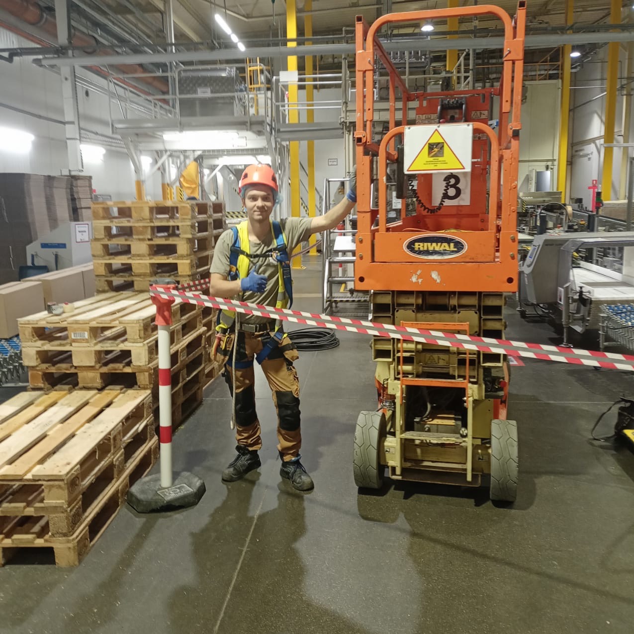 Pracownik w kasku i uprzęży stoi obok podnośnika koszowego Riwala w hali produkcyjnej, zabezpieczony taśmą ostrzegawczą. W tle palety i instalacje.