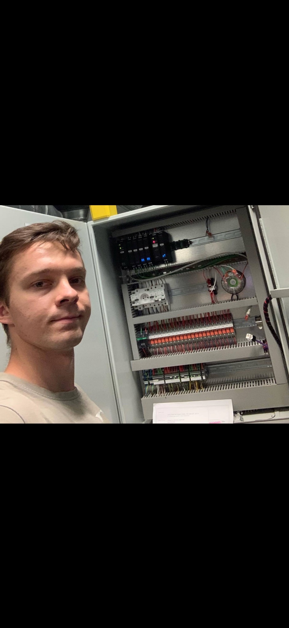 Młody elektryk prezentuje rozdzielnicę elektryczną z licznymi przewodami, bezpiecznikami i transformatorem, dokumentacja techniczna na dole, oświetlenie LED w tle.