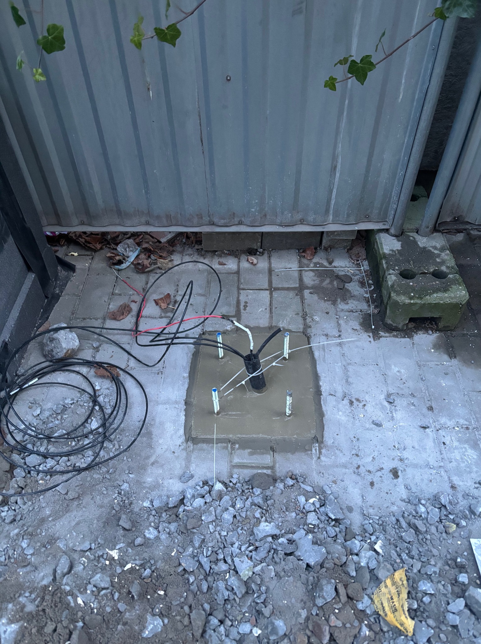 Fundament pod instalację odgromową z wyprowadzonymi przewodami i uziomami, widok z góry na betonową podstawę i kable. Tło stanowi metalowe ogrodzenie i fragment roślinności.