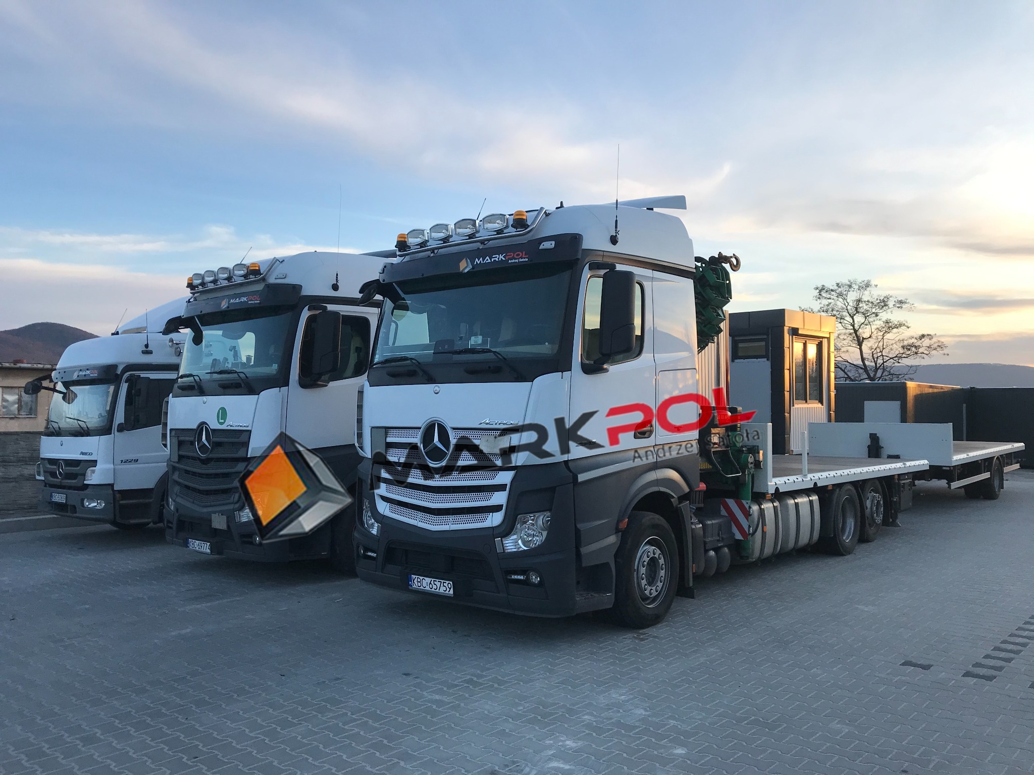 Trzy białe ciężarówki Mercedes-Benz z platformami transportowymi, zaparkowane na brukowanym placu, z widocznym zachodem słońca w tle i logo firmy 'MARKPOL' na kabinie.