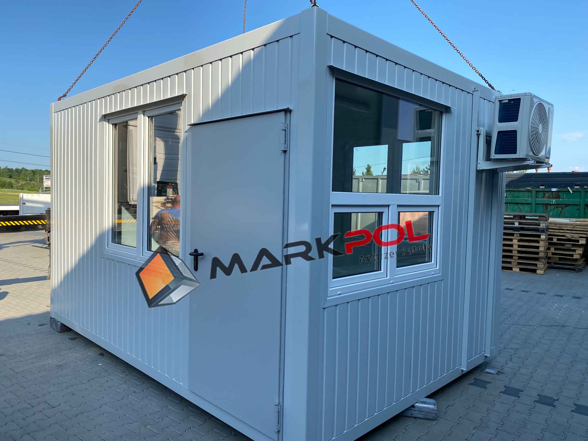 Szary kontener biurowy z klimatyzacją, podnoszony za pomocą łańcuchów, z widocznym logo firmy MARKPOL na ścianie.