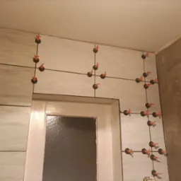 Układanie jasnych, marmurkowych płytek w łazience z użyciem systemu poziomowania, widoczne krzyżyki dystansowe i kliny, surowa ściana w tle.