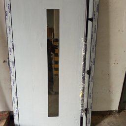 KasMar MariuszPorzeziński - Białe drzwi z podłużnym przeszkleniem, obramowane czarną ramą z logo Alufrof, zabezpieczone folią. Widoczne elementy montażowe i odbicie osoby w szybie.