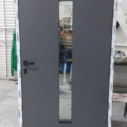 KasMar MariuszPorzeziński - Szare drzwi z szybą, obramowane profilem Alupfrof, widoczne w warsztacie. Drzwi z czarną klamką, ustawione pionowo, gotowe do montażu. Widoczne narzędzia i elementy montażowe.