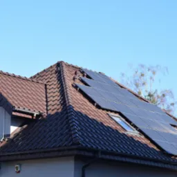 Brązowy dach z dachówki ceramicznej z zamontowanymi panelami słonecznymi, widoczne okna dachowe i klimatyzator na ścianie budynku.