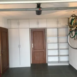 Garaż z zabudową meblową: białe szafki wiszące i stojące, drewniane drzwi, puste białe regały, rower zawieszony na ścianie, szara podłoga z płytek.