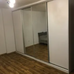 Nowoczesna szafa przesuwna z lustrzanymi drzwiami, białe fronty, widoczna w lustrze część pokoju z szarą sofą, drewniana podłoga.