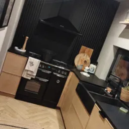 Nowoczesna kuchnia z jasnymi drewnianymi szafkami, czarnym blatem, piekarnikiem retro i czarną ścianą z pionowych paneli, uzupełniona dywanem w geometryczne wzory.