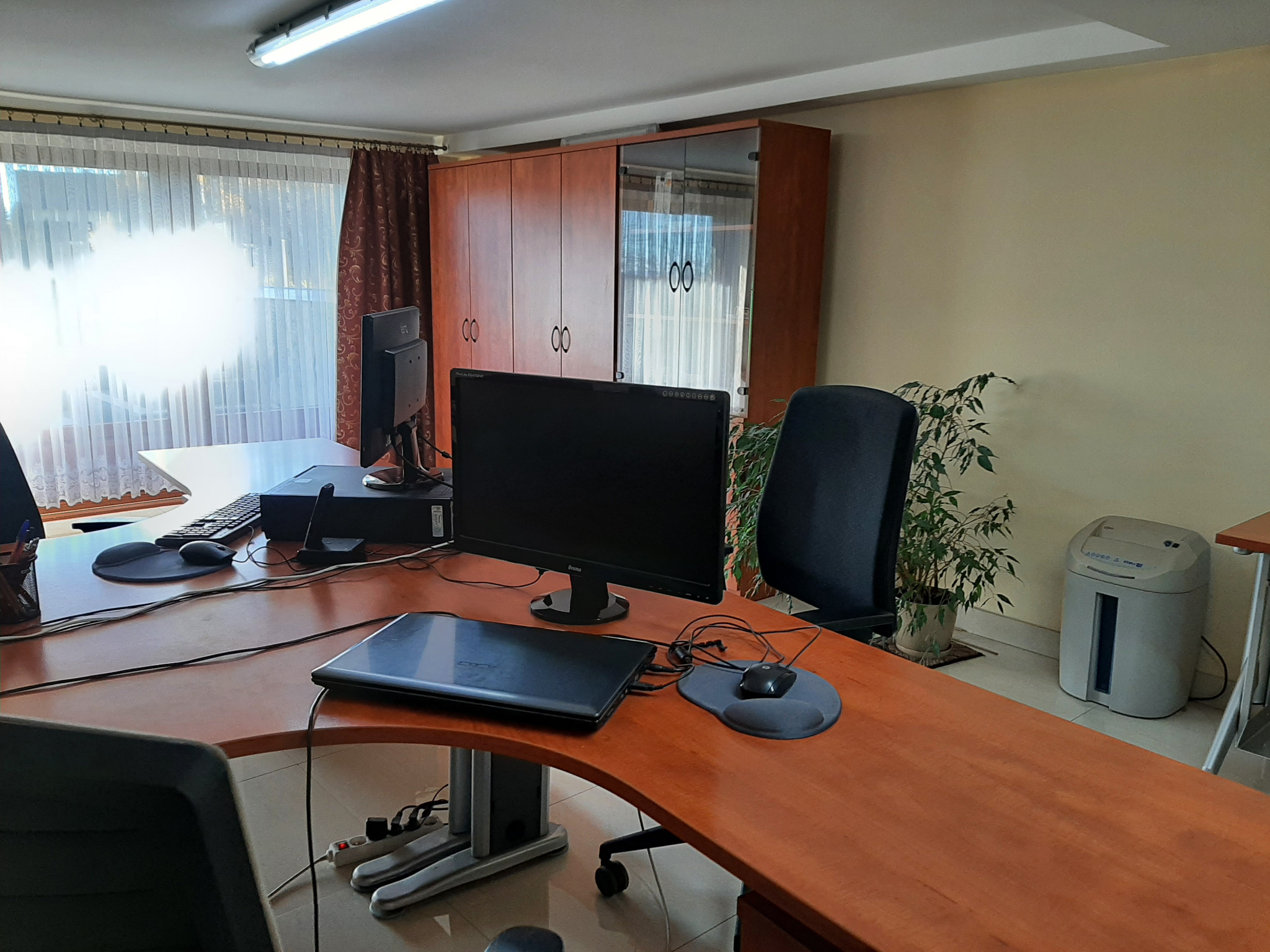 Biuro z biurkami, komputerami i szafą w tle. Na biurku laptop, myszka i podkładka. W rogu pokoju roślina doniczkowa i niszczarka do dokumentów.