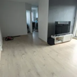 Jasne panele podłogowe w salonie z pomalowaną na szaro ścianą, widoczny odkurzacz i wejście do kuchni.