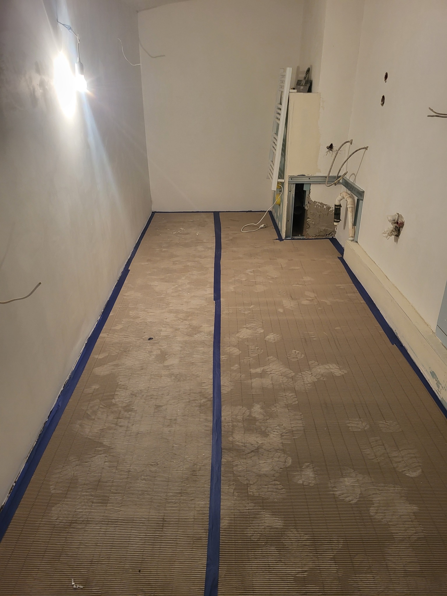Przygotowanie podłoża pod wylewkę w pomieszczeniu. Mata izolacyjna rozłożona na podłodze, zabezpieczona niebieską taśmą malarską przy ścianach. Widoczne instalacje.