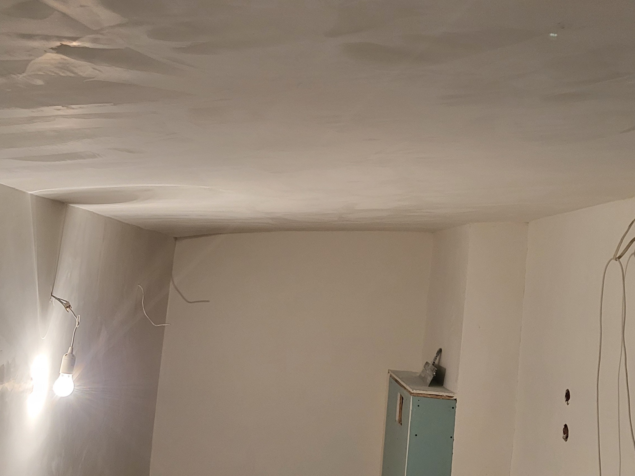Pokój po nałożeniu gładzi na ścianach i suficie. Widoczne niedoskonałości powierzchni i prowizoryczne oświetlenie żarówką. Mały fragment szafki w rogu pokoju.