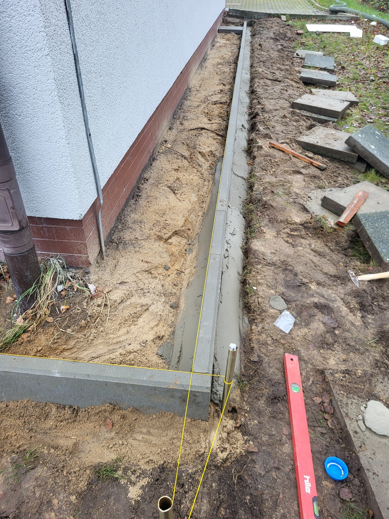 Układanie obrzeży betonowych przy budynku, widoczne narzędzia i poziomica. Obrzeża wyznaczone żółtym sznurkiem, etap prac przygotowawczych do układania kostki.