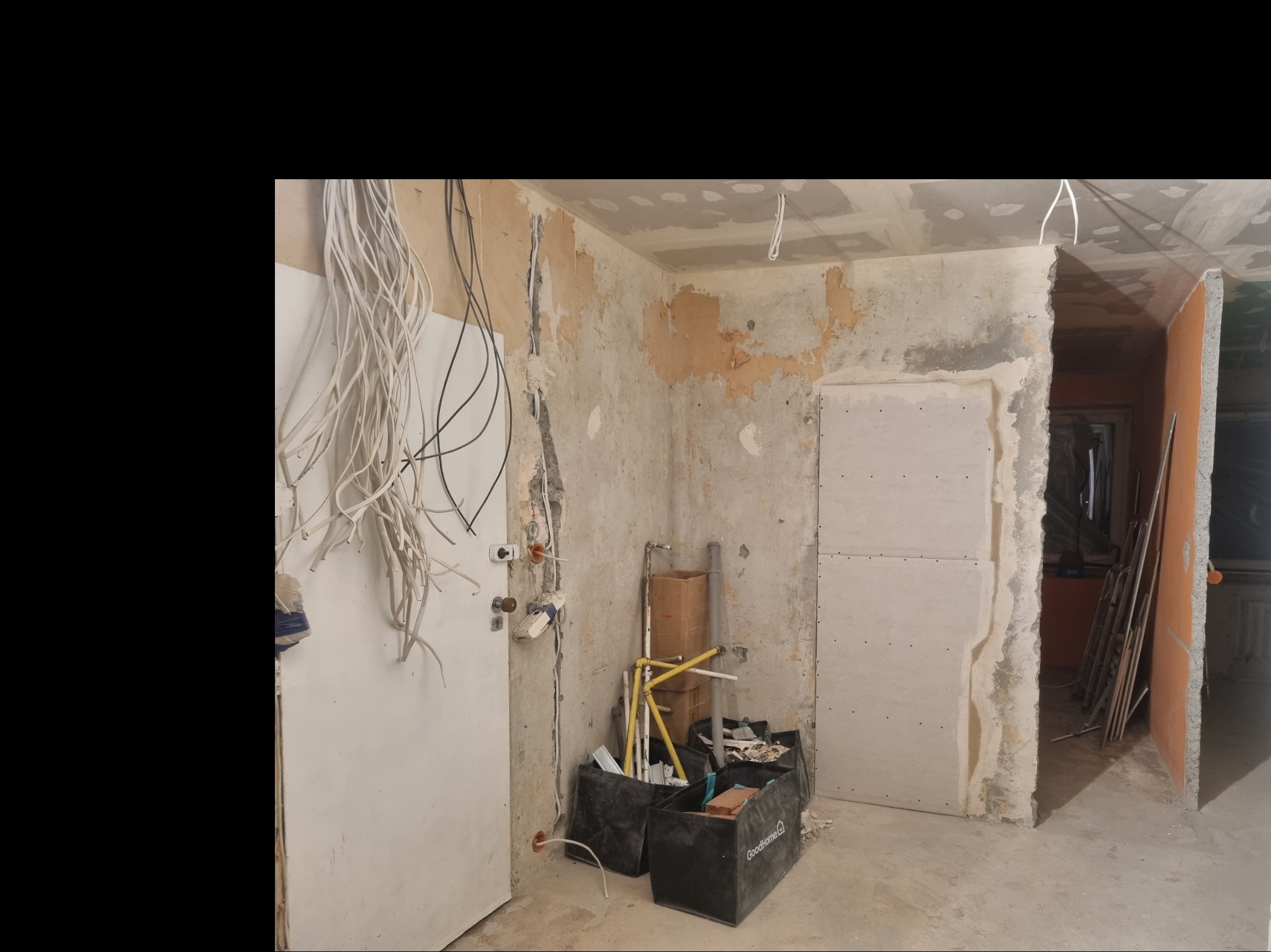 Wnętrze w trakcie remontu: odsłonięte przewody elektryczne, fragmenty starej farby na ścianach, płyty gipsowo-kartonowe, narzędzia w czarnych pojemnikach, widok na sąsiednie pomieszczenie...