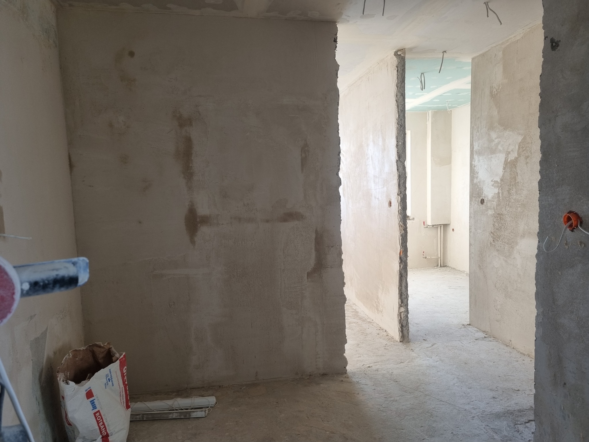 Wnętrze w trakcie prac wykończeniowych: surowe ściany z widocznymi śladami szpachlowania, odsłonięte przewody elektryczne, worek z zaprawą Knauf Rotband i narzędzie do gładziowania w rogu...