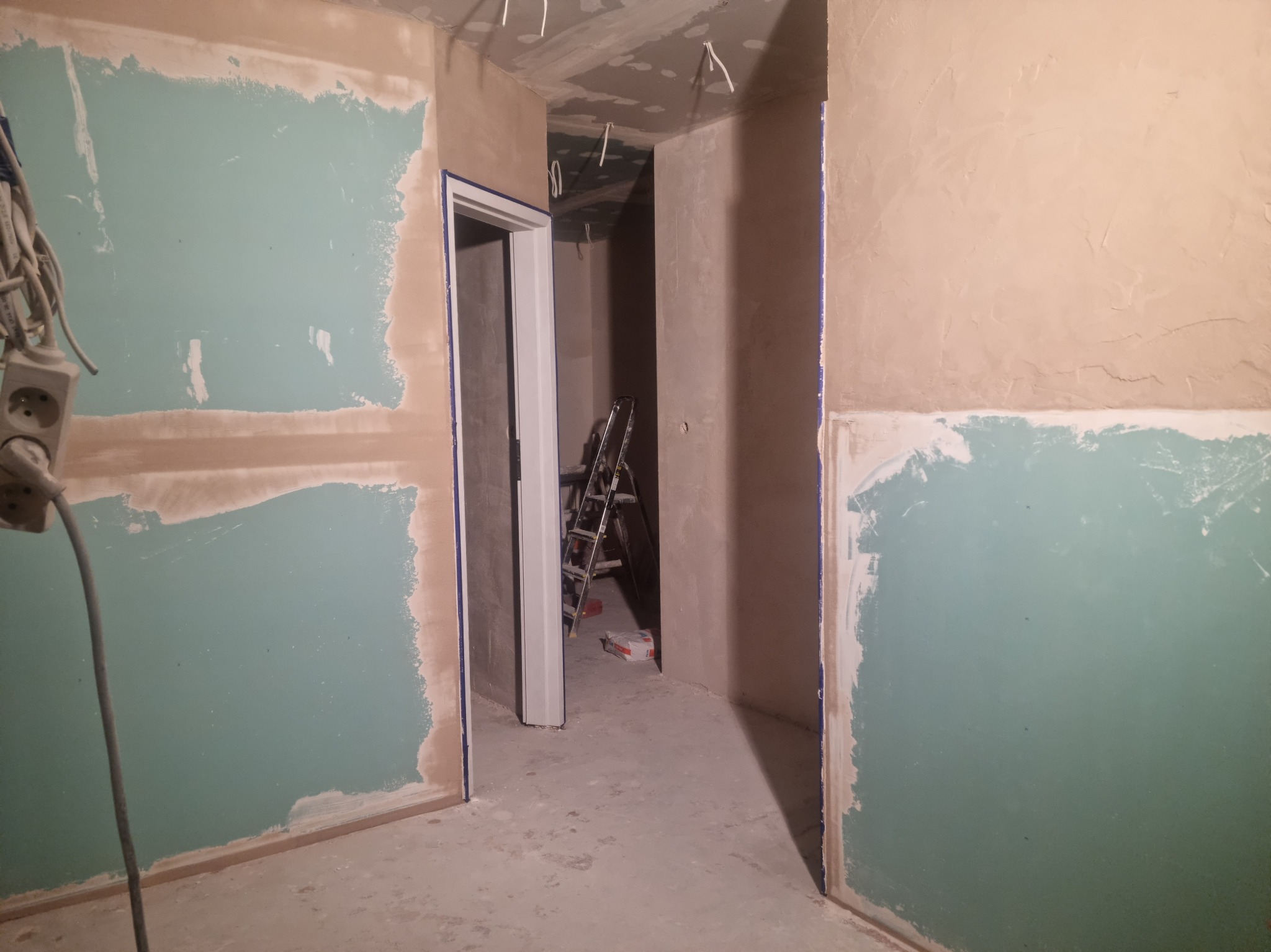 Wnętrze w trakcie remontu: ściany częściowo pokryte zieloną farbą, widoczne niedoskonałości i nierówności, przewody elektryczne wystające z sufitu, drabina w głębi pomieszczenia, surowy stan...