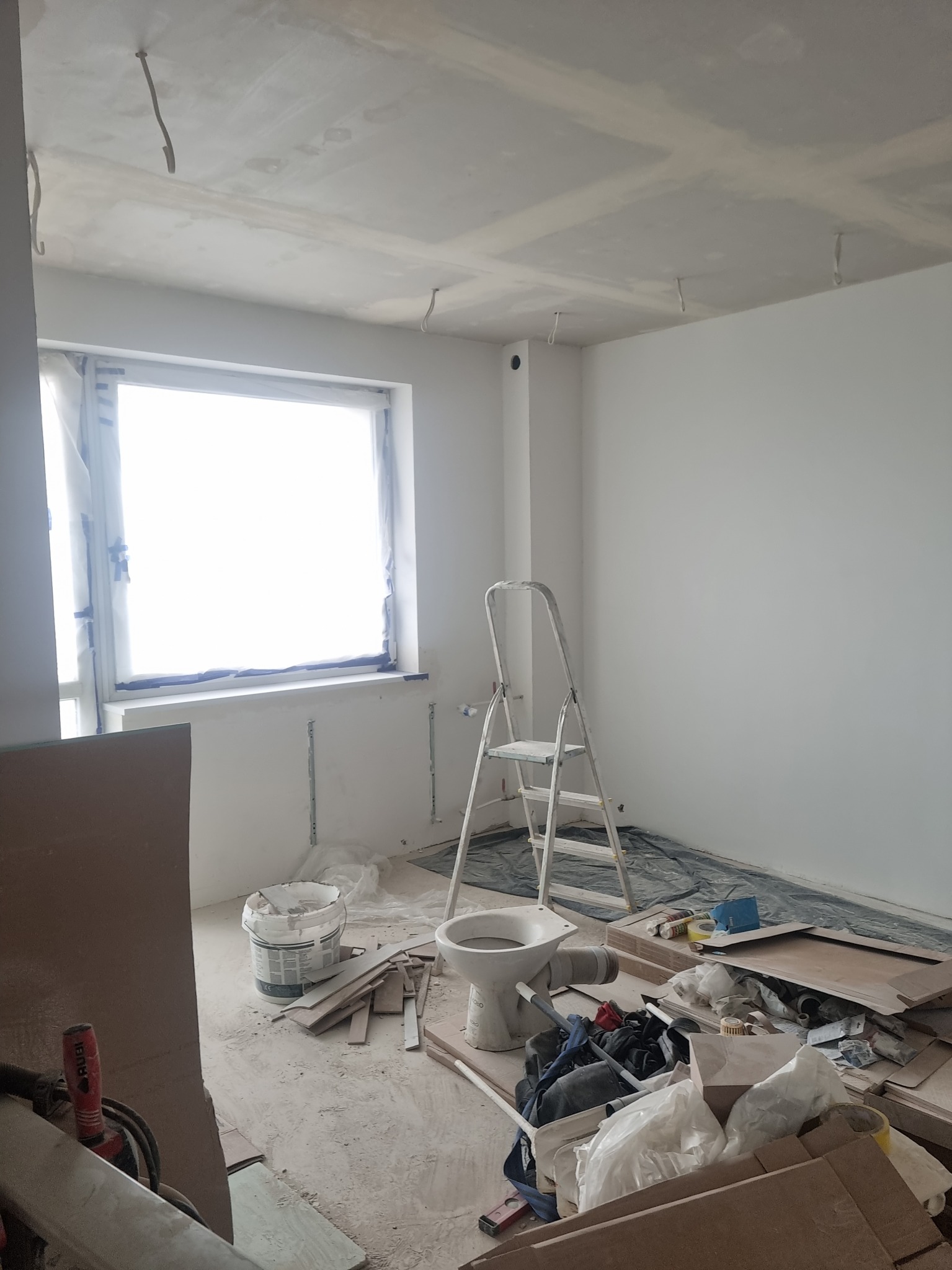 Wnętrze w trakcie remontu: świeżo pomalowane białe ściany, surowy sufit z wystającymi kablami, okno zabezpieczone folią, drabina oparta o ścianę, na podłodze resztki materiałów budowlanych i stare WC.