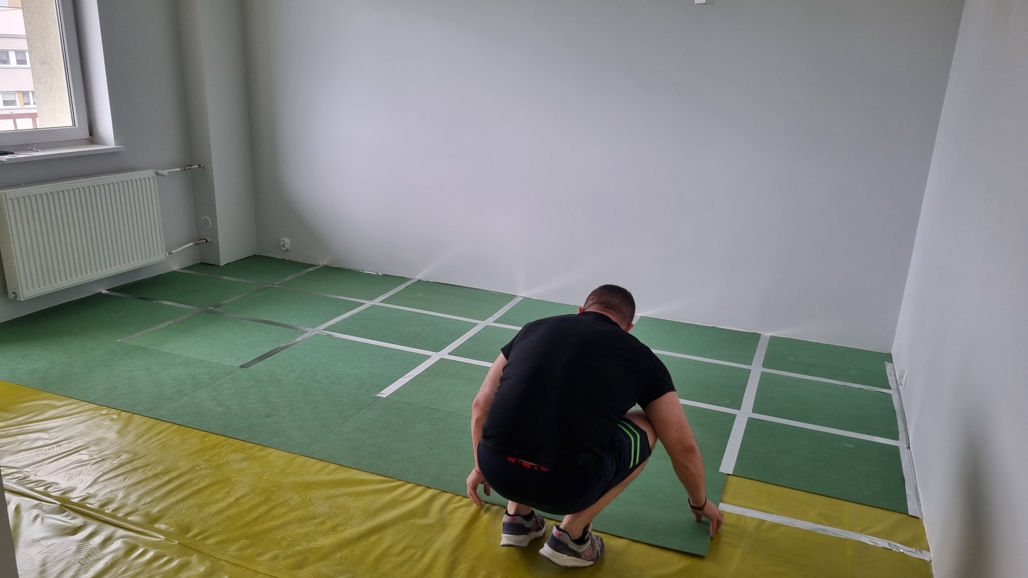 Mężczyzna w czarnej koszulce i krótkich spodenkach układa zielone płyty podłogowe na żółtej folii w pustym, jasnym pokoju z grzejnikiem pod oknem.