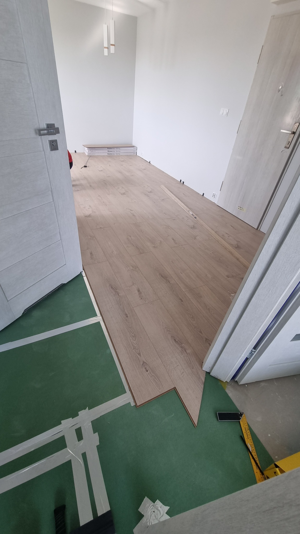 Fragment układania jasnych paneli podłogowych w pomieszczeniu, widoczna zielona podkładka z taśmą, drzwi wejściowe i narzędzia pomiarowe.