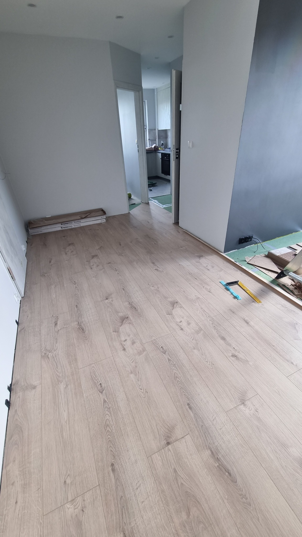 Jasne panele podłogowe ułożone w nowym mieszkaniu, widoczne narzędzia pomiarowe i paczki paneli przy ścianie, ściany białe i szara, w tle widoczna kuchnia.