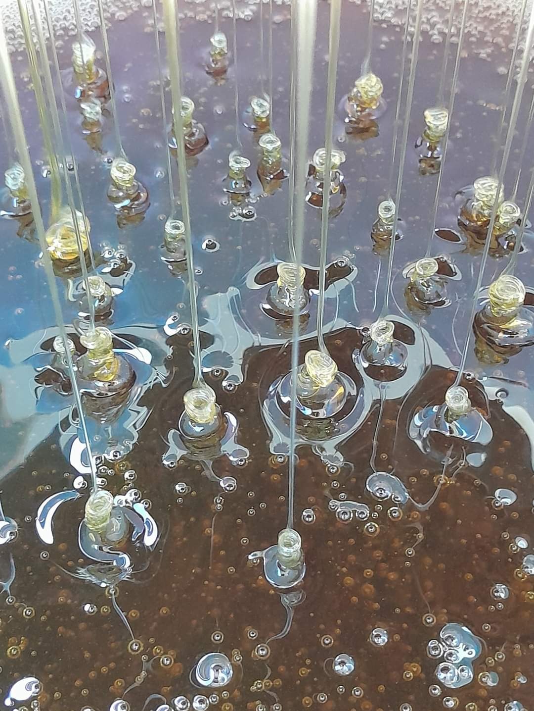 Strumienie gęstej, ciemnej cieczy opadają na lustrzaną powierzchnię, tworząc liczne, małe kopułki i pęcherzyki powietrza.