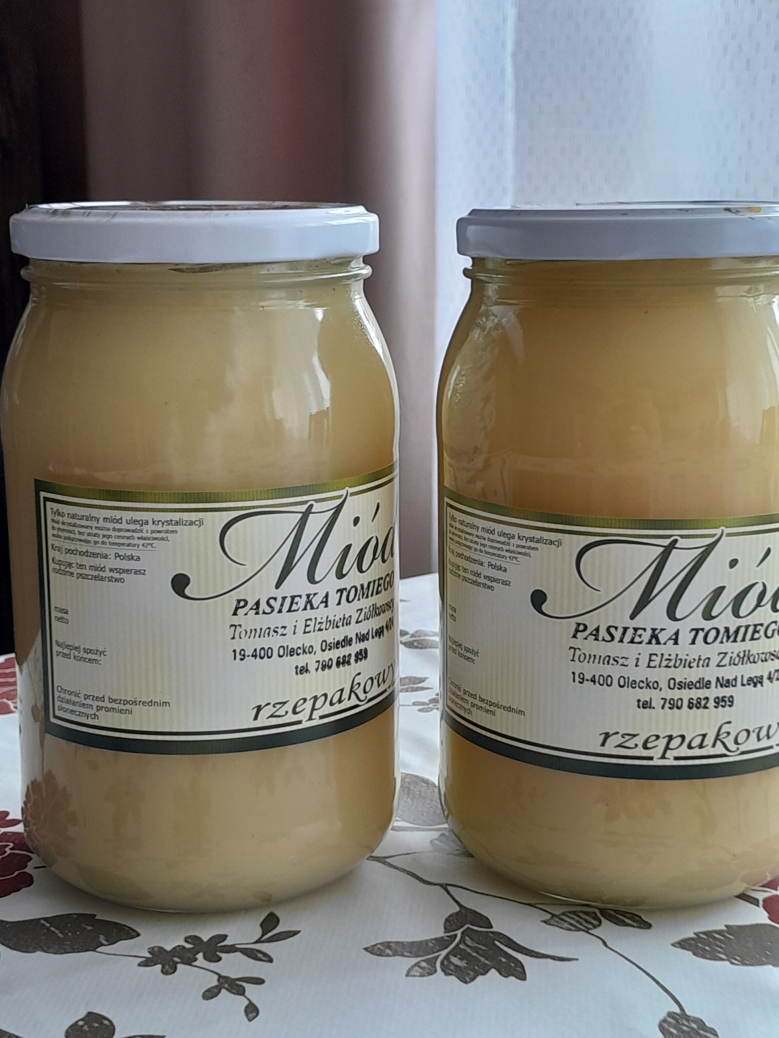 Dwa słoiki z jasnym miodem rzepakowym, etykiety z nazwą pasieki Tomiego, widoczne na kwiecistym obrusie.