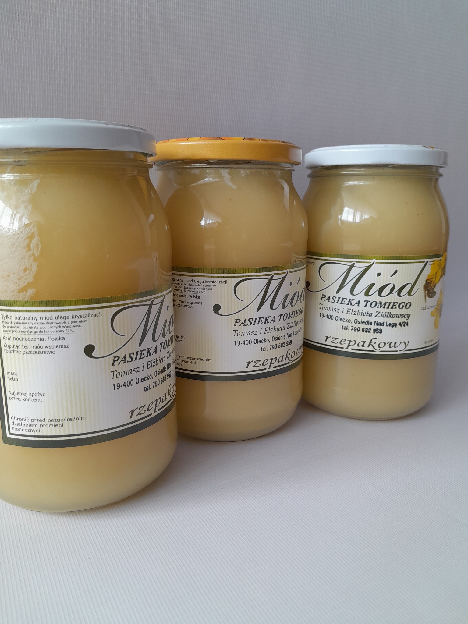Trzy słoiki z miodem rzepakowym od Pasieki Tomiego, ustawione na białym tle, z widocznymi etykietami i różnymi kolorami wieczek.