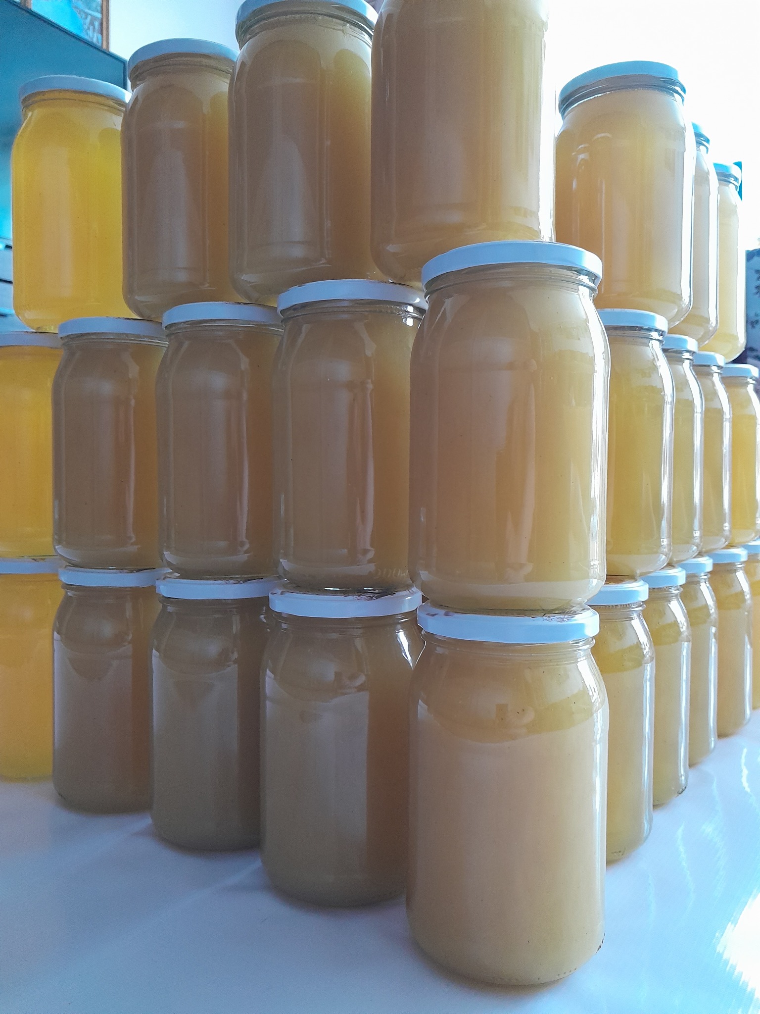 Ułożone piramidalnie słoiki z miodem o różnej barwie, od jasnożółtej do ciemniejszej, z białymi zakrętkami, tworzące ekspozycję.