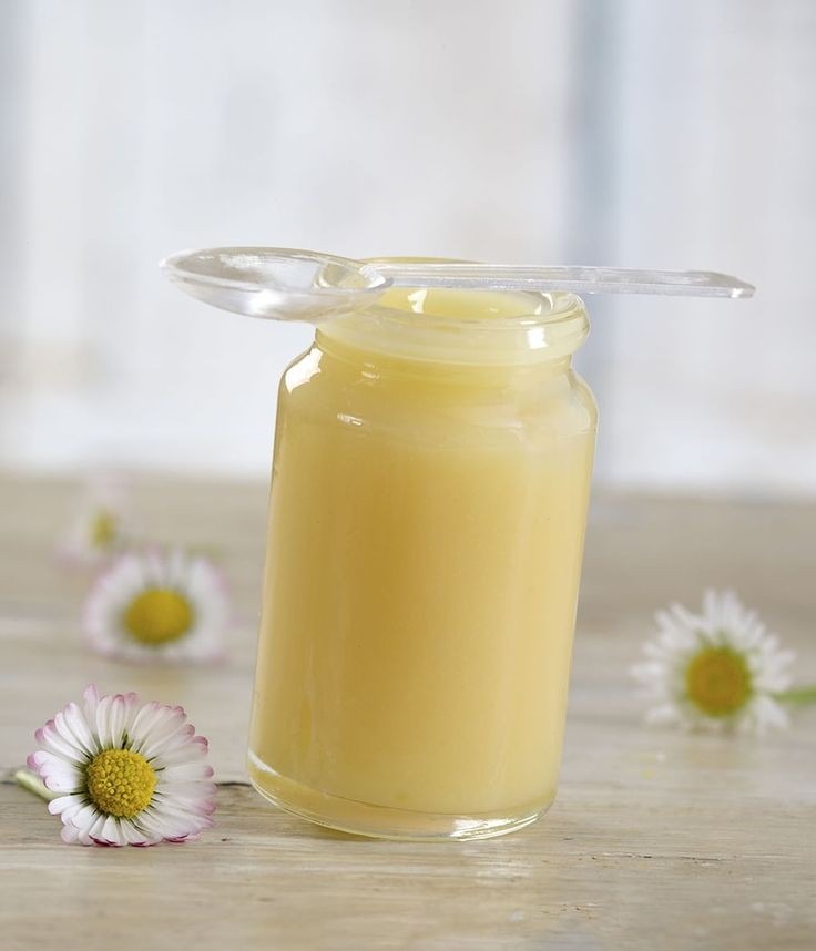 Słoik z miodem kremowanym, przezroczysta łyżeczka na wierzchu, trzy stokrotki w tle i na pierwszym planie, drewniany blat.