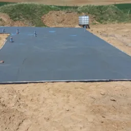 Wylana płyta betonowa, przygotowana pod budowę domu, z wystającymi rurami instalacyjnymi, otoczona piaskiem i ziemią.