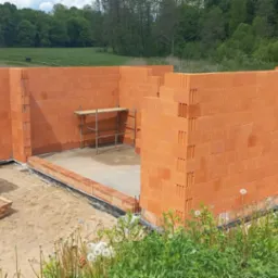 Wznoszone ściany parteru domu z czerwonej cegły ceramicznej na fundamencie, widoczne rusztowanie wewnątrz, palety z bloczkami i deskami obok.