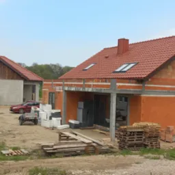Dwa domy w stanie surowym otwartym: jeden z murowanymi ścianami z czerwonej cegły i czerwoną dachówką, drugi otynkowany z drewnianą elewacją szczytową, na placu budowy z widocznym materiałem...