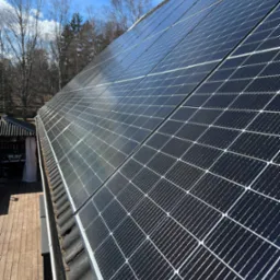 Panel słoneczny na dachu domu z widocznymi zabrudzeniami, rynna z liśćmi, drewniany taras i błękitne niebo z drzewami w tle.