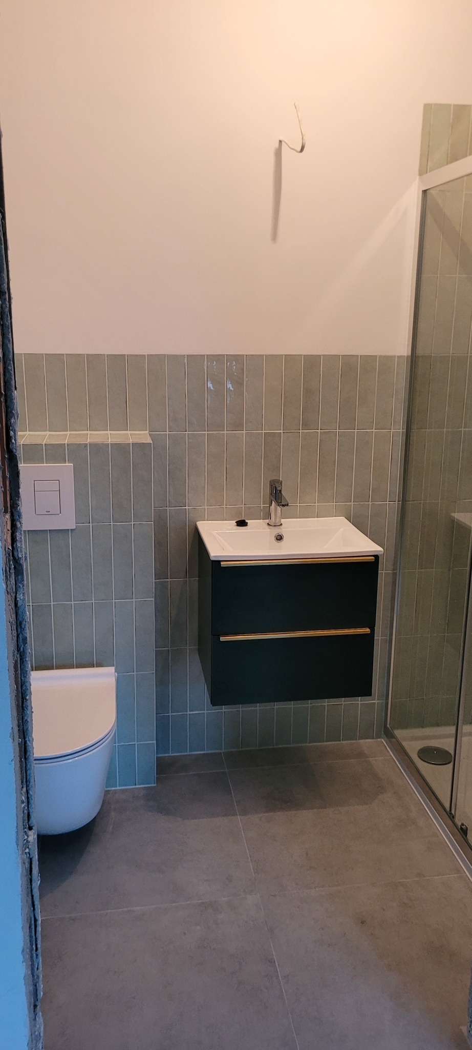 Wykończona łazienka z wiszącą szafką umywalkową w kolorze butelkowej zieleni z złotymi uchwytami, płytkami w odcieniach zieleni ułożonymi pionowo, toaletą w rogu i kabiną prysznicową z szklanymi...