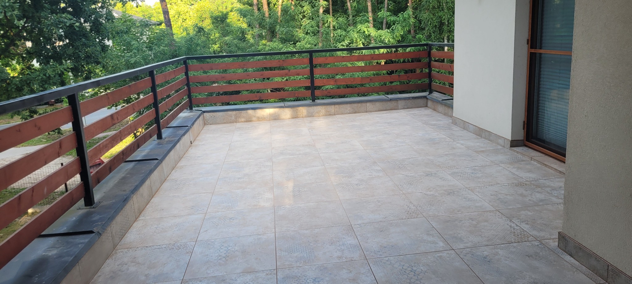 Wykończony balkon z beżowymi płytkami podłogowymi o wzorzystej powierzchni i drewnianą balustradą z czarnymi metalowymi elementami, widok na zieleń drzew.